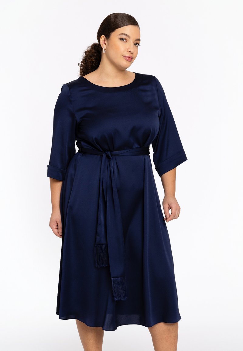 Navy Satin-Kleid mit drei Viertel-Ärmeln, tailliertem Schnitt und Gürtel mit Quastenakzenten, fließendem Rock, der unter das Knie fällt.