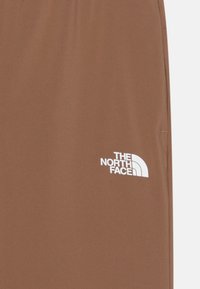 Shorts de sport marron en tissu lisse, avec un logo blanc sur le côté gauche. Le design comprend une taille élastique et des coutures latérales.