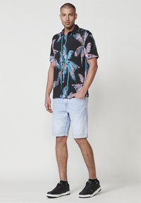 Camisa negra de manga corta con un colorido patrón de palmeras, combinada con pantalones cortos de mezclilla claros y zapatillas negras.