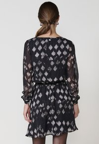 Schwarzes, gemustertes Kleid mit langen Ärmeln, aus einem strukturierten Stoff und mit einem elastischen Bund. Die Rückseite hat einen Knopfverschluss und einen fließenden Saum.