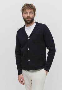 Donker marineblauwe cardigan van textuurstof, voorzien van een gelabelde kraag, twee voorzakken en witte knopen. Draag het over een wit shirt.