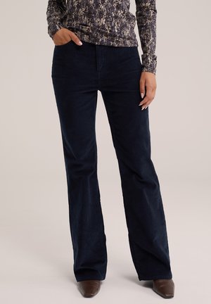 Persona con pantalones de pana azul oscuro, una camiseta de manga larga estampada y zapatos marrones de punta afilada, con una mano en el bolsillo.