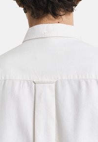 Camicia bianca in cotone con colletto a punta, chiusura con bottoni al collo e dettaglio a pieghe sul retro. Tessuto liscio e leggero.