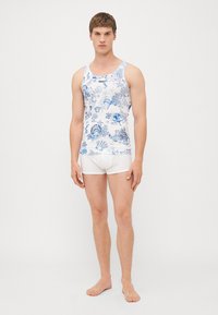 Weißes Tanktop mit blauem meeresthematischem Muster, versehen mit blauen Akzenten und einem Logo am Halsauschnitt; kombiniert mit weißen Shorts.