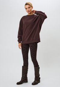 Sudadera grande marrón, leggings negros con estampado de leopardo y botas altas marrones oscuras con detalles de herrajes y hebillas.