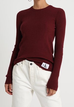 Mujer con un ajustado top de manga larga acanalado color granate, metido dentro de unos pantalones blancos de talle alto, mostrando una etiqueta visible con el logo de Calvin Klein.