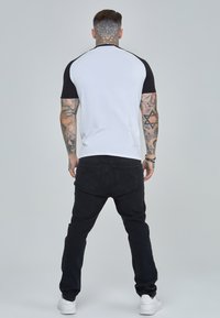 Camiseta de manga corta en blanco y negro con un diseño raglán, combinada con pantalones vaqueros negros y zapatillas blancas, mostrando tatuajes en los brazos.