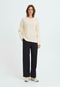 Krämfärgad stickad tröja med nedhasade axlar och ribbat nederkant, tillsammans med svarta vida byxor och beige sneakers mot en neutral bakgrund.