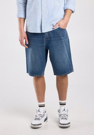 Man draagt een blauw gestreept overhemd, losse denim shorts, witte sokken met zwarte strepen en witte Air Jordan sneakers, staand tegen een effen achtergrond.