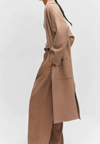 Lange beige cardigan van zacht materiaal, met een losse pasvorm, wijde mouwen en een voorzak, gecombineerd met een hoge taille bruine broek.