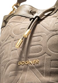Ein taupefarbenes Wildleder-Bucketbag mit geprägtem Logo-Detail. Es verfügt über metallische graue Akzente mit goldfarbenen Beschlägen und einem verstellbaren Kordelzugverschluss.
