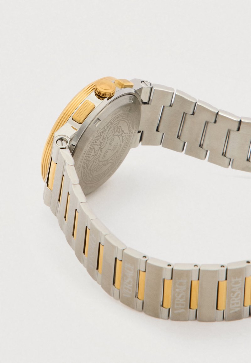 Silber- und goldfarbene Metall-Armbanduhr mit strukturiertem Gehäuse und Armband, ausgestattet mit einer gemusterten Rückseite und gravierten Logoakzenten auf den Gliedern.