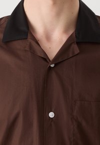 Gros plan d'une personne portant une chemise marron à boutons avec un col noir et une poche de poitrine visible.