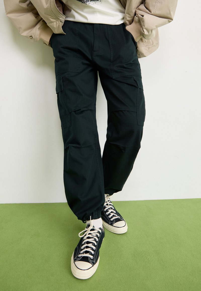 Pantalons cargo noirs avec une finition texturée, dotés de poches latérales et de poignets élastiques, associés à des baskets noires et à une veste beige.