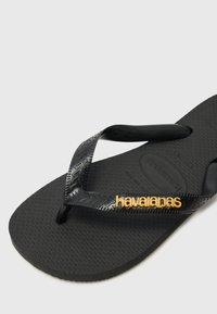Black rubber flip-flop with textured sole and black straps, featuring gold "havaianas" logo on the strap.