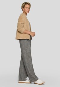 Frau steht in einer beigen, flauschigen Strickjacke, grauen karriert-weiten Hosen und weißen Sneakern vor einem schlichten grauen Hintergrund.