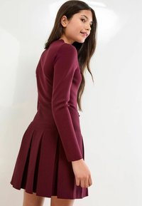 Burgundy színű, hosszú ujjú ruha, amelyet egy testhez simuló felsőrész és bővülő, redőzött szoknya jellemez. Simára kidolgozott, enyhe fényű textúrával.