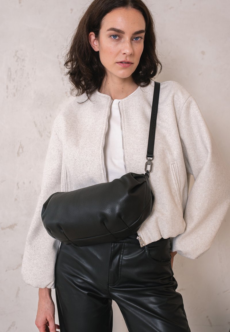 Schwarze Leder-Crossbody-Tasche mit runder Form und Reißverschluss, kombiniert mit einer hellgrauen, strukturierten Bomberjacke und schwarzen Hosen.