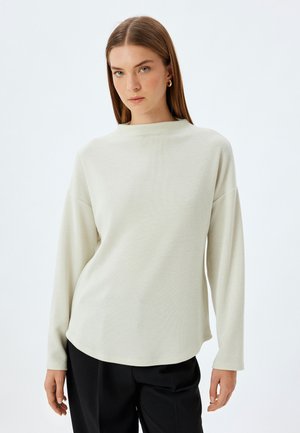 Haut beige clair à manches longues avec un motif texturé, encolure ronde et coupe décontractée ; associé à un pantalon noir.