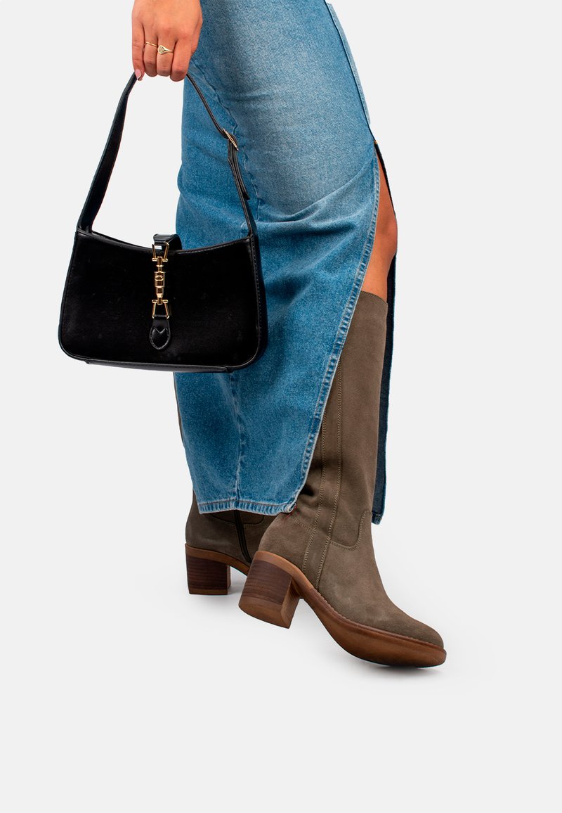 Bottes en cuir suédé vert olive, montantes jusqu'au genou, avec un talon bloc, assorties à un sac à main noir brillant avec un fermoir et une bandoulière dorés. Jupe en denim avec une fente.