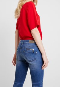 Top corto de manga corta rojo combinado con jeans de mezclilla azul que presentan bolsillos traseros, detalles de cremallera y un degradado de lavado claro.