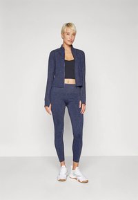 Tamsiai mėlynas sportinis rinkinys, kuriame yra aptempta, tekstūruota striukė su užtrauktuku, derančios tamprės ir juodas trumpas marškinėlis, papildytas baltais sportbačiais.