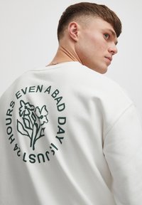 Hvid sweatshirt med rund halsudskæring med grøn blomsterdesign og tekst på ryggen: "24 TIMER SELV EN DÅRLIG DAG ER BARE."