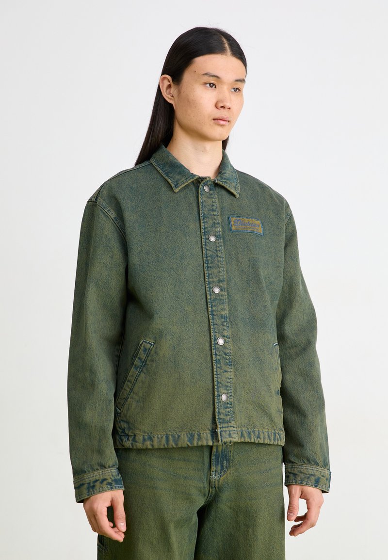 Jachetă din denim verde cu guler clasic, nasturi frontali cu capse și buzunare laterale. Prezintă un finisaj texturat și un mic patch cu logo.