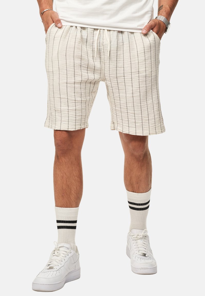 Helle beigen Shorts mit vertikalen Streifen, elastischem Bund und Taschen. Kombiniert mit weißen Turnschuhen und gestreiften weißen Socken.