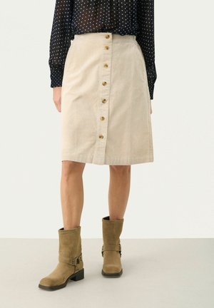 Beige Cord-Rock aus Kord mit einer vorderen Knopfleiste und A-Linien-Silhouette. Kombiniert mit braunen Stiefeletten und einem gepunkteten Blusenhemd.