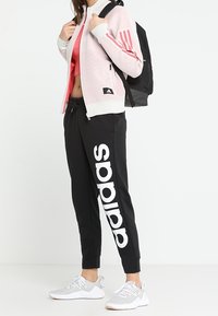 Chaqueta rosa y blanca con cremallera y tres rayas rojas en las mangas, pantalones de chándal negros con un gran logo blanco de "adidas" y zapatillas deportivas grises.