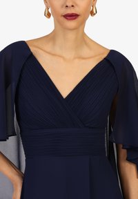 Marineblaues Kleid mit gefaltetem Oberteil und V-Ausschnitt, featuring transparente Umhangärmel und einem figurbetonten Taillendesign. Goldene Ohrringe runden den Look ab.