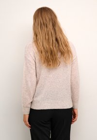 Frau mit langen welligen blonden Haaren, die einen hellbeigen Pullover und schwarze Hosen trägt, steht vor einer schlichten weißen Wand.