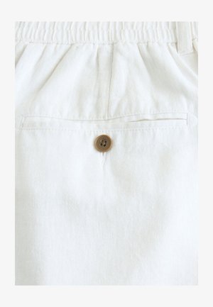 Läheltä kuvattu valkoinen pellavakangas, jossa on takatasku, joka on kiinnitetty ruskealla napilla, korostaen sen rakennetta ja yksinkertaista muotoilua.