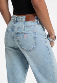 Jeans de tiro alto en azul claro con una textura ligeramente desgastada, que presentan una etiqueta de cuero y un logo triangular de la marca en el bolsillo trasero.