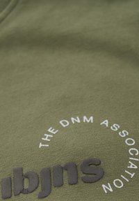 T-shirt en coton olive, avec un design circulaire de texte blanc et des lettres noires en relief, texture lisse avec un tissu côtelé.