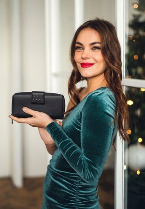 Black leather wallet with a zip closure, accented by a metallic clasp featuring studs. Held by a person in a teal velvet dress.