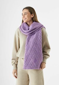 Vrouw die glimlacht, gekleed in beige loungewear en een grote geribbelde paarse sjaal om haar nek gewikkeld, staande tegen een witte achtergrond.