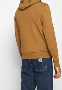 Sweat à capuche marron avec ourlet et poignets côtelés, associé à un pantalon en denim bleu comportant un patch logo sur la poche arrière.