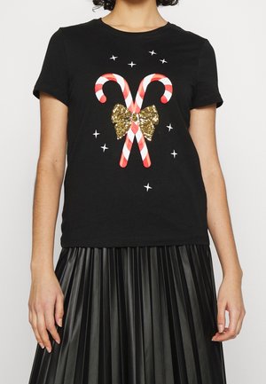 Kvinde iført sort T-shirt med krydsede rød- og hvide slikstokke, guldglitrende sløjfe og omgivende hvide stjerneformer.