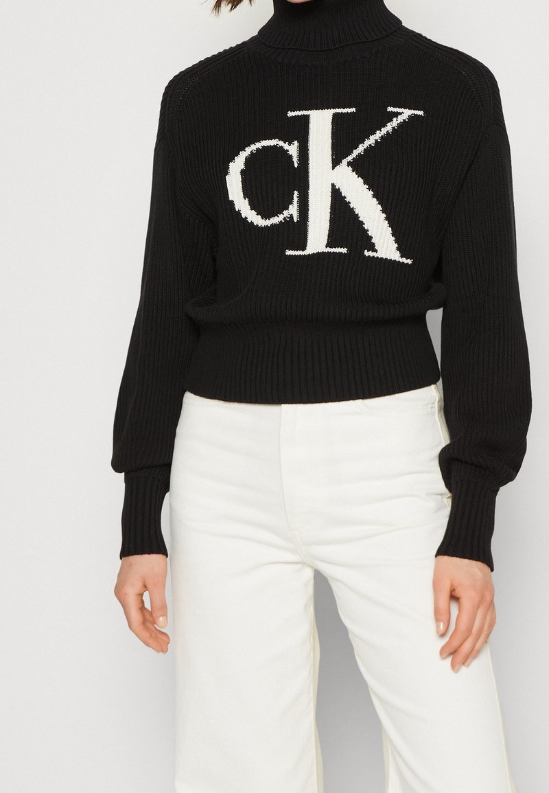 Pull à col roulé côtelé noir avec grand logo blanc « cK » associé à un pantalon blanc taille haute, montré du cou aux hanches.