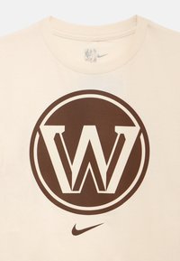 Krémové tričko z bavlny s velkým hnědým kruhem, ve kterém je bílé logo "W", a pod ním malým logem Nike. Klasický střih s kulatým výstřihem.