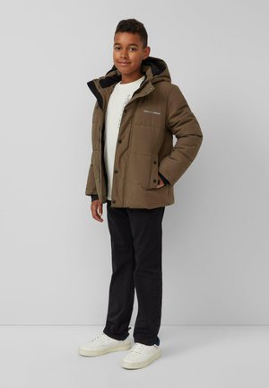 Braune gepolsterte Jacke mit Kapuze, ausgestattet mit schwarzen Akzenten und Knöpfen, getragen über einem weißen Hemd und schwarzen Hosen, kombiniert mit weißen Schuhen.
