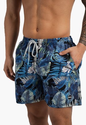 Torse d'homme portant un short de bain bleu à imprimé feuilles tropicales avec un cordon blanc, main dans la poche et tatouage tribal sur l’avant-bras droit.