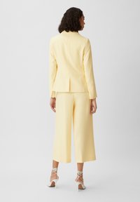 Yellow tailored suit with a structured blazer and wide-leg trousers. Smooth fabric, minimalist design, paired with white strappy heels.