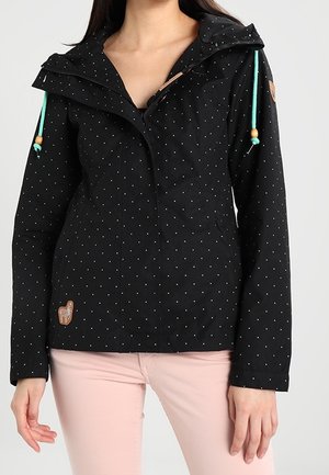 Frau trägt eine schwarze, langärmelige Jacke mit kleinen weißen Punkten und türkisen Kordeln, kombiniert mit hellrosa Hose.