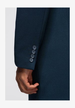 Marineblauwe textuurjas mouw met vier bijpassende knopen en een getailleerde pasvorm, met een hand gepositioneerd nabij de manchette.