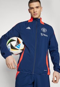 Giacca sportiva blu navy con accenti rossi, dotata di zip completa, logo sul petto e polsini elastici. Il modello tiene in mano un pallone da calcio.