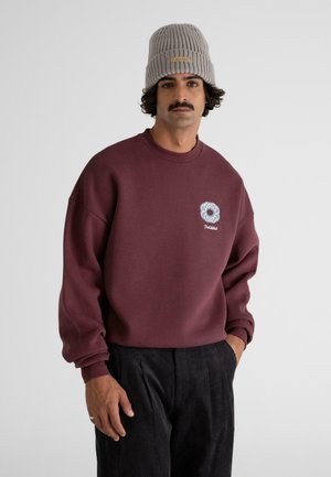 Maroon sweatshirt made of cotton, featuring a crew neck, loose sleeves, and a light blue embroidered logo on the chest. Worn with a gray knit hat.