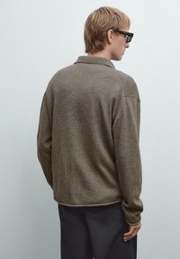 Brun stickad tröja med krage, strukturerad tyg, avslappnad passform och ribbade mudd. Modellen visas bakifrån mot en ljus bakgrund.